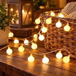 Puede incluir: Una guirnalda de luces LED con bombillas esféricas de color blanco cálido sobre un cable fino, dispuesta sobre una superficie de madera frente a una cesta de mimbre trenzado y un farol. Las pequeñas bombillas esmeriladas emiten luz suave.