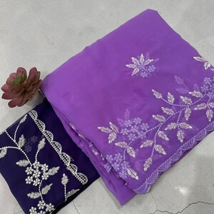 Peut inclure: Un morceau de tissu violet plié avec une broderie florale argentée sur le bord, associé à une section de tissu bleu foncé plus petite présentant une bordure en dentelle festonnée blanche et des motifs de broderie florale argentée.