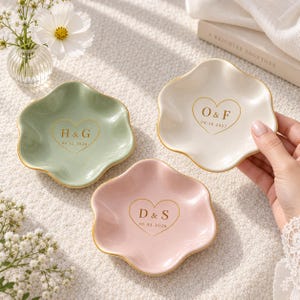 Könnte beinhalten: Drei gewellte Keramikschalen in Salbeigrün, Creme und zartem Rosa. Jede Schale hat einen goldfarbenen Rand und ein goldenes Herzmotiv in der Mitte mit Initialen und Daten: H & G 06.12.2026, O & F 04.18.2027 und D & S 10.03.2026.
