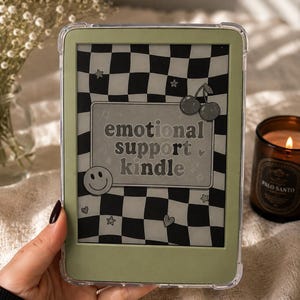 Peut inclure: Une liseuse électronique dotée d'un cadre vert sauge et d'une coque de protection transparente. L'écran affiche un fond à damier noir et blanc avec une étiquette rectangulaire centrale portant le texte emotional support kindle, des cerises, un smiley et des étoiles.