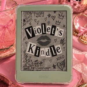 May include: A light green e-reader device encased in a clear protective cover. The screen displays a gray background with black hand-drawn doodles of hearts, stars, and the text "READ MORE" and "XOXO" surrounding the words "Violet's Kindle".