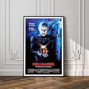 Puede incluir: Un póster de película Hellraiser enmarcado que muestra una figura pálida con alfileres en la cabeza, vestida de cuero negro, sosteniendo una caja de rompecabezas brillante. El texto dice: Demon to some. Angel to others. HELLRAISER. He'll tear your soul apart.