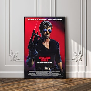 May include: A framed film poster for Cobra featuring Sylvester Stallone wearing blue-tinted sunglasses and holding a submachine gun with a laser sight. The background is a vibrant red with the text Crime is a disease. Meet the cure. and Stallone Cobra.