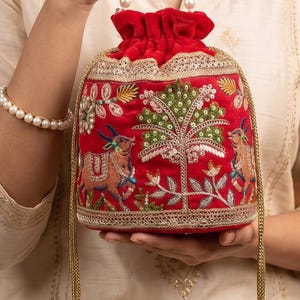 May include: A red velvet drawstring pouch bag featuring intricate embroidery of animals and a central tree motif. The bag is accented with gold metallic trim and a handle made of white faux pearls, with matching pearl tassels hanging from the drawstrings.