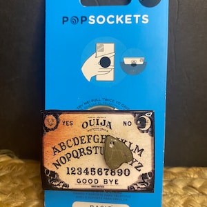 Op de afbeelding: Een zwart-wit Ouija-bord met een groene planchette erop. Het bord heeft de letters van het alfabet, de cijfers 1-9 en 0, en de woorden "YES", "NO" en "GOOD BYE". Het bord bevindt zich op een blauwe achtergrond met de woorden "BASIC" en "POPSOCKETS" erop.