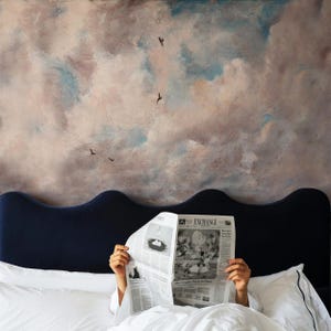 May include: A dark blue wavy headboard sits against a wall mural featuring painted clouds in shades of beige, blue, and gray with small birds. A person holds an open newspaper titled Exchange in front of their face while in bed.