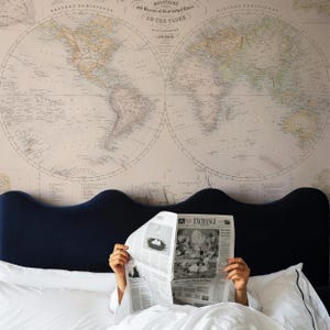 May include: A person reading a newspaper titled Exchange whilst lying in a bed with a dark blue scalloped headboard. The wall behind the bed features a large, vintage-style world map print with beige tones and detailed geographical labels.