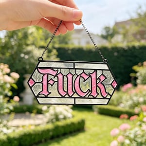 May include: A hexagonal stained glass suncatcher featuring the word FUCK in bold pink lettering with a black outline. The background consists of textured clear glass panels framed in black metal, suspended by a thin black metal chain.