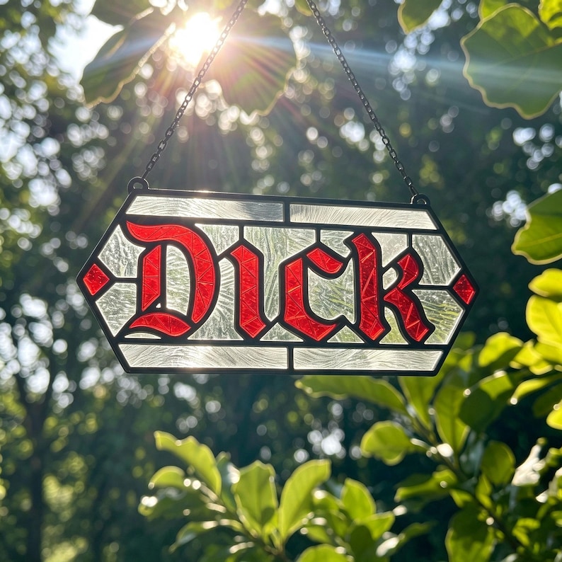 May include: A hexagonal stained glass suncatcher featuring the word "Dick" in bold, red, textured glass letters. The background consists of clear, rippled glass panels framed in black metal, suspended by a thin black chain for hanging display.