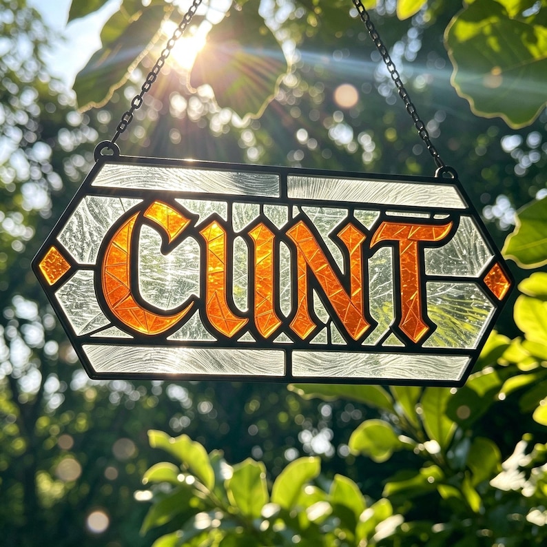 May include: A rectangular stained glass suncatcher featuring the word CUNT in bold orange lettering. The letters are set against a clear, textured glass background with a black metal frame and a hanging chain attached to the top corners.