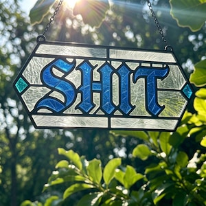May include: A hexagonal stained glass suncatcher featuring the word SHIT in bold blue gothic lettering. The letters are set against a background of textured clear glass, framed by a black metal border with small teal diamond accents on the sides.