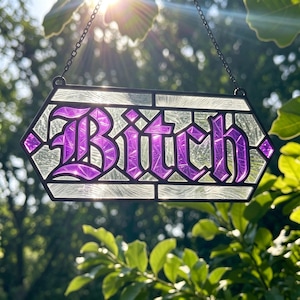 May include: A hexagonal stained glass hanging sign featuring the word Bitch in bold purple lettering with a textured finish. The piece is framed in black metal and suspended by a thin black chain, allowing light to pass through the glass panels.