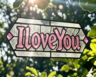 May include: A hexagonal stained glass suncatcher featuring the text I Love You in pink, glittery lettering. The piece is framed in black metal with clear, textured glass panels surrounding the words and small pink diamond accents on the sides.