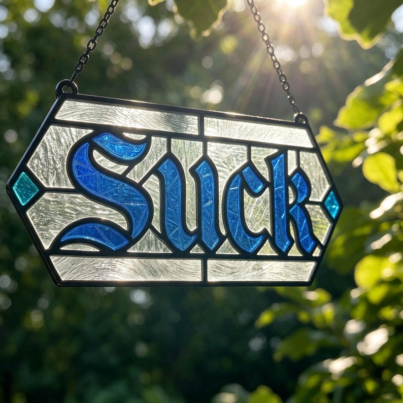 May include: A hexagonal stained glass suncatcher featuring the word SUCK in bold blue glass letters against a textured clear glass background. The piece is framed in black metal and hangs from a black metal chain, catching the sunlight.