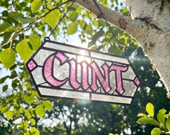May include: A hexagonal stained glass suncatcher featuring the word CUNT in bold pink lettering. The piece is constructed with black metal lead lines and textured clear glass panels, suspended by a thin black metal chain for hanging.