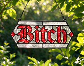 May include: A hexagonal stained glass suncatcher featuring the word Bitch in bold red gothic lettering. The letters are set against a background of textured clear glass, all encased in a black metal frame with a hanging chain attached.