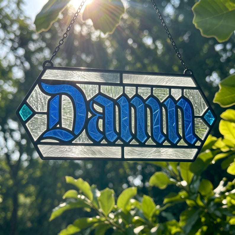 May include: A hexagonal stained glass suncatcher featuring the word "Damn" in bold blue gothic lettering. The piece is constructed with textured clear glass panels and framed in black metal, suspended by a thin black chain for hanging.