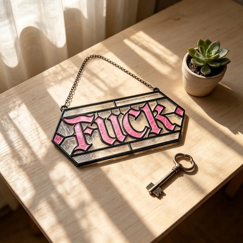 May include: A stained glass hanging sign with a black metal frame and a chain, featuring the word FUCK in pink lettering against a clear, textured glass background. Beside the sign lies a vintage-style metal skeleton key with a dark finish.
