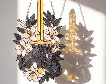 May include: A stained glass suncatcher featuring a central yellow sword held by two dark gray armored gauntlets, surrounded by white lily flowers and dark gray leaves. The piece is suspended by a metal chain and casts a shadow on the wall.