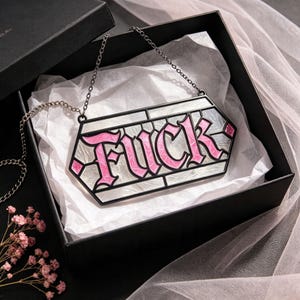 May include: A hexagonal pendant necklace featuring the word FUCK in pink lettering with a white marbled background, enclosed in a black metal frame. The piece hangs from a thin black chain and is presented inside a black gift box with white tissue.