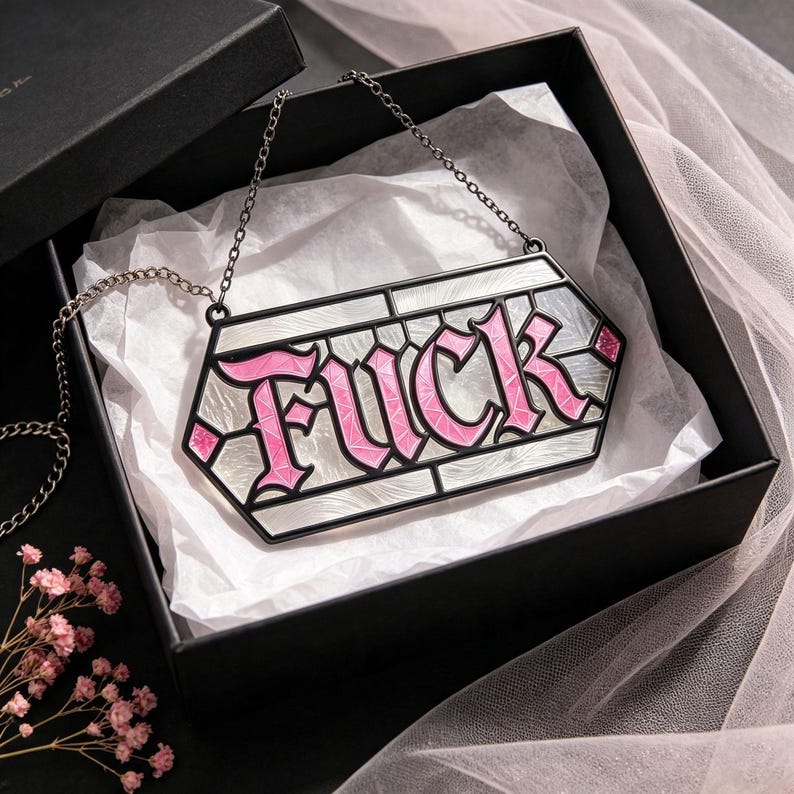 May include: A hexagonal pendant necklace featuring the word FUCK in pink lettering with a white marbled background, enclosed in a black metal frame. The piece hangs from a thin black chain and is presented inside a black gift box with white tissue.
