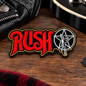 Peut inclure: Un écusson brodé présentant le texte rouge RUSH et un logo starman blanc sur fond noir avec une bordure jaune. À côté se trouve un médiator noir avec le texte blanc ROCK NEVER DIES et une petite étoile imprimés sur la surface.