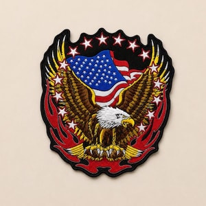 May include: An embroidered patch featuring a bald eagle with brown and white feathers, positioned in front of a waving American flag. The design is framed by red flame-like shapes and a circular arrangement of white stars on a black background.