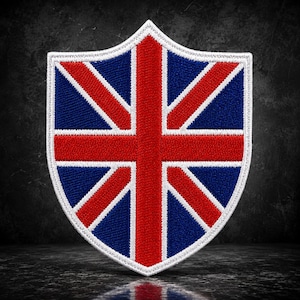 Puede incluir: Un parche bordado con forma de escudo que presenta el diseño de la bandera Union Jack. El parche muestra una cruz roja central y franjas diagonales rojas delineadas en blanco sobre un fondo azul oscuro, bordeado por un grueso borde cosido en blanco.