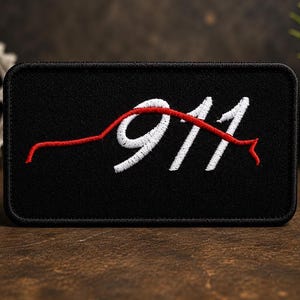 May include: A rectangular black embroidered patch featuring the white stitched text 911. A continuous red stitched line forms the silhouette of a sports car profile that curves over the numbers, creating a distinct graphic design on the fabric.