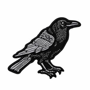May include: An embroidered iron-on patch featuring a black and white raven in profile. The bird is depicted with detailed stitching that highlights its feathers, beak, and talons, creating a high-contrast graphic design on a solid background.