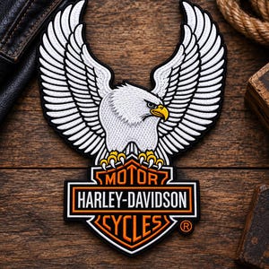 May include: An embroidered patch featuring a white eagle with spread wings, yellow talons, and a yellow beak, positioned above the orange, black, and white Harley-Davidson Motor Cycles logo with the registered trademark symbol in the bottom right corner.