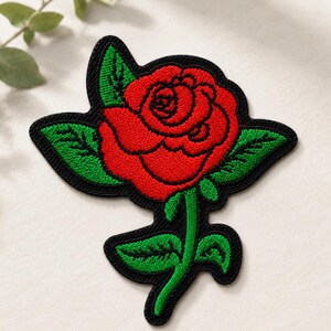May include: An embroidered patch featuring a vibrant red rose with detailed green leaves and a stem, all outlined in a bold black border. The design is stitched with dense thread, showcasing a classic floral motif on a black fabric backing.