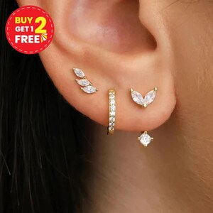 Peut inclure: Un ensemble de boucles d'oreilles dorées comprenant un clou incurvé avec trois pierres claires taille marquise, une petite créole pavée de pierres, un clou en forme de V avec deux pierres taille marquise et un clou rond avec une pierre claire.