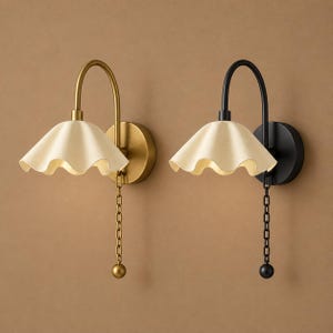 May include: Two wall sconces featuring curved arms and ruffled, off-white shades. One sconce has a brushed brass finish with a matching chain pull, while the other has a matte black finish with a matching chain pull and spherical end.