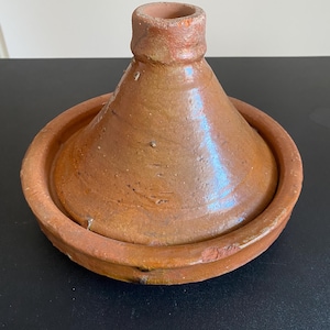Può includere: Un tajine in terracotta marrone composto da una base circolare larga con un bordo basso e un coperchio conico alto con una piccola apertura sulla parte superiore. La superficie dell'utensile in argilla ha una finitura lucida e smaltata.