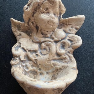 Può includere: Un'acquasantiera da parete testurizzata di colore beige raffigurante un cherubino scolpito con ali. L'oggetto ha un aspetto simile alla pietra invecchiata con accenti marrone scuro nelle fessure, che mettono in risalto i dettagli in rilievo.