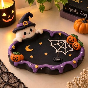 Può includere: Un vassoio ovale nero con bordo smerlato viola, decorato con un fantasma bianco che indossa un cappello da strega nero, tre zucche arancioni, una ragnatela bianca, un piccolo ragno nero, una luna crescente gialla e diverse stelle gialle.