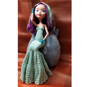 May include: A fashion doll with purple and green hair, wearing a mint green crochet mermaid gown and blue gloves. The doll is posed on a grey plush pillow. The dress features a strapless bodice and a flared, openwork skirt.