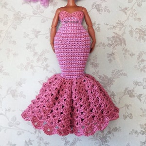 May include: A pink crocheted mermaid-style dress for a doll. The dress has a fitted bodice and a flowing skirt with a layered design.