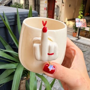Puede incluir: Una taza de cerámica color crema con un asa esculpida en tres dimensiones que representa una mano haciendo un gesto de dedo medio. La mano tiene una uña pintada de rojo y un pequeño corazón rojo en la punta. Una cadena dorada con un pequeño dije de estrella iridiscente cuelga del dedo.