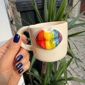 Puede incluir: Una taza de cerámica de color crema que presenta un diseño esculpido en tres dimensiones de un par de labios en la parte frontal. Los labios están pintados con rayas verticales en una paleta de colores arcoíris, incluyendo rojo, naranja, amarillo, verde, azul y morado.