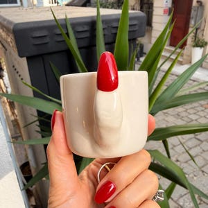 Puede incluir: Una taza de cerámica de color crema con un asa en forma de dedo humano con una uña pintada de rojo brillante. La taza tiene un cuerpo cilíndrico liso y un tono neutro sólido, lo que aporta un elemento táctil único al diseño del objeto.