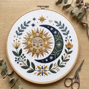 May include: A circular embroidery hoop containing white fabric with a hand-stitched design of a central sun with a face, a dark blue crescent moon, and surrounding botanical elements including green leaves, small blue berries, and gold flowers.