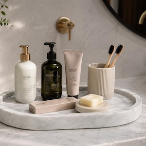 May include: A white marble oval tray holds a collection of bathroom essentials: a white lotion bottle with a gold pump, a dark green soap dispenser with a black pump, a beige tube, a beige ribbed cup with two bamboo toothbrushes, and a bar of soap.