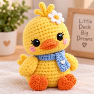 May include: A yellow crocheted duck toy featuring a bright orange beak and feet, large black safety eyes, and pink blush cheeks. The duck wears a light blue crocheted scarf with a small white heart button and a white flower on its head.