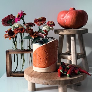 Può includere: Un vaso cilindrico in ceramica con un rilievo a foglia arancione testurizzato, posizionato su un rialzo rotondo in legno. Vicino si trovano un vaso in legno con provette e fiori di zinnia, uno sgabello, una zucca rossa e peperoncini rossi.