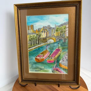 May include: A framed watercolor painting depicting a canal scene with two colorful barges floating on green water. In the background, stone buildings and a bridge are visible under a blue sky. The artwork is enclosed in a textured gold frame.