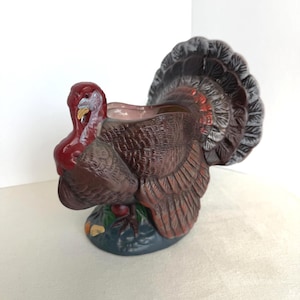 May include: A ceramic turkey-shaped gravy boat or planter featuring a brown body with textured feather details, a red wattle, and a fanned tail. The base is dark blue with green accents, and the interior of the vessel is a light pinkish hue.