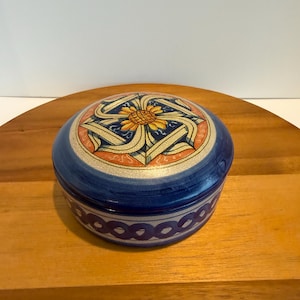 May include: A round ceramic lidded box featuring a crackled glaze finish. The lid displays a central yellow sunflower motif surrounded by blue and cream geometric patterns. The base is decorated with a repeating dark blue circular chain design.