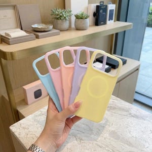 May include: A set of five silicone smartphone cases in pastel colors including light blue, pink, peach, lavender, and yellow. Each case features a large rectangular camera cutout and a circular magnetic charging ring on the back surface.